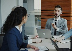 A professional consultant speaks to a client while both are seated at a desk in an office.